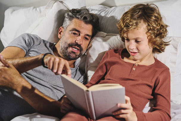 Man in sleepwear helping boy read interesting book while resting in bedroom at home - Photo, Image