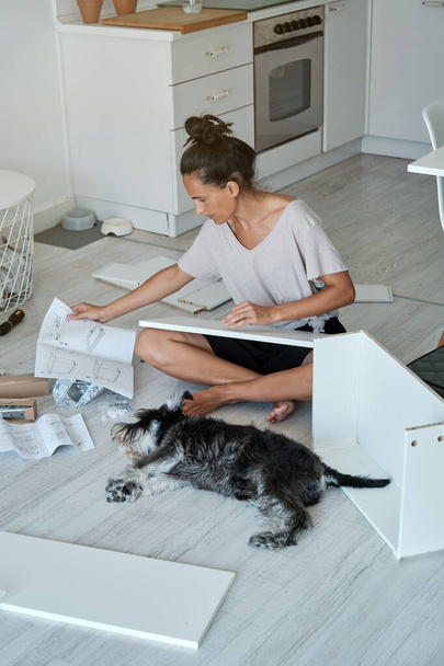 High angle full length of concentrated woman with hair bun sitting on floor with crossed legs and reading manual while assembling furniture in kitchen with fluffy Miniature Schnauzer - Fotografie, Obrázek