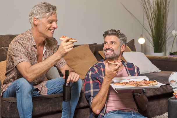 Délicieux hommes adultes en vêtements décontractés manger une pizza appétissante tout en s'amusant dans le salon confortable à la maison - Photo, image