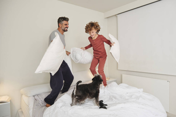 Full body of cheerful adult father and curly haired son in pajamas jumping on bed with pillows while playing with dog - Photo, Image