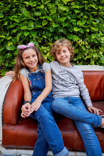 Glückliche preteen Jungen und Mädchen in lässiger Kleidung lächeln und schauen in die Kamera, während sie auf einer alten Ledercouch in der Nähe von grünen Büschen im Garten sitzen - Foto, Bild
