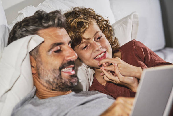 Dorosły mężczyzna i nastoletni syn w piżamie czytają razem ciekawą bajkę na dobranoc i uśmiechają się wieczorem w sypialni w domu - Zdjęcie, obraz