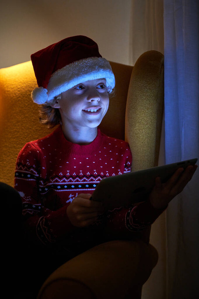 Charmant garçon en chapeau Santa et pull en utilisant une tablette tout en étant assis sur un fauteuil confortable et en regardant ailleurs - Photo, image