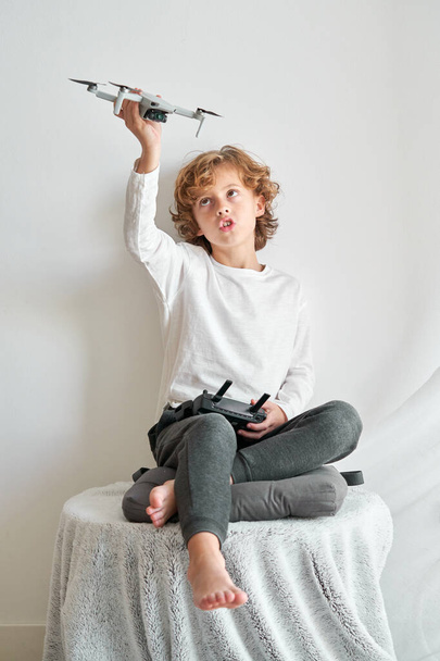 Lunghezza completa del bambino biondo seduto sul tavolo e guardando in alto mentre si diverte con il drone giocattolo - Foto, immagini