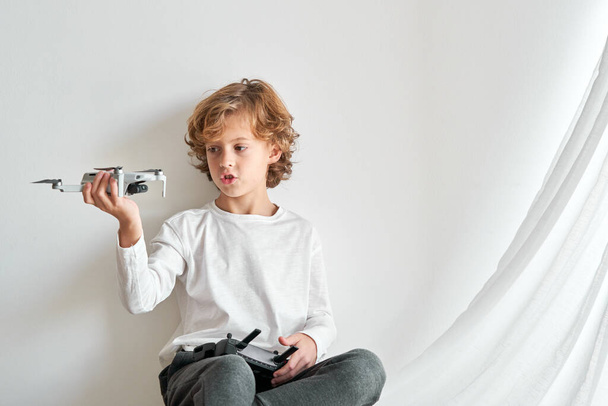 Geïsoleerd schattig krullend kind zittend tegen een witte achtergrond en kijkend naar speelgoed drone in de hand - Foto, afbeelding