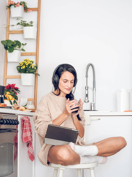 Corpo completo di donna ispanica concentrata in cuffia seduta con tazza di caffè e tablet di navigazione in cucina - Foto, immagini