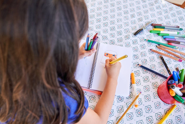 Van bovenaf bekijken van creatief kind zitten aan tafel en tekening afbeelding in notitieblok met kleurrijke potloden overdag - Foto, afbeelding