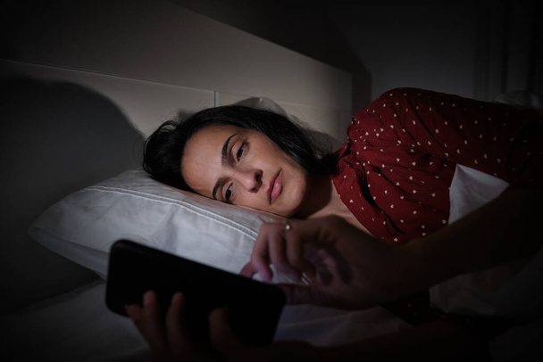 Mujer hispana enfocada en la mensajería de texto de ropa de dormir en el teléfono celular moderno mientras está acostada en una cómoda cama tarde en la noche en el dormitorio - Foto, imagen