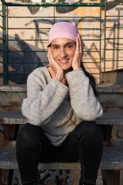 Очаровательная взрослая латиноамериканка в розовом платке символизирует международную кампанию по борьбе с раком молочной железы, сидящая на гранже, трогая лицо и глядя в камеру - Фото, изображение