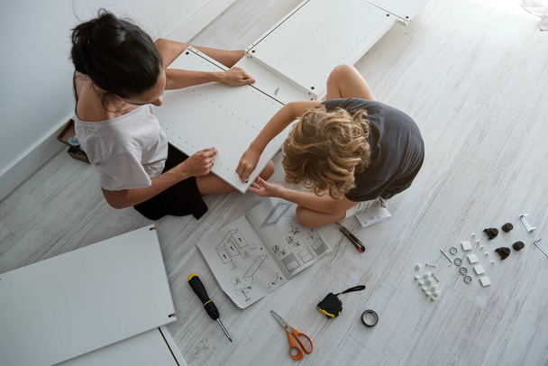 Top view of unrecognizable mother and son in domestic clothes sitting on floor near manual and assembling wooden wardrobe with various instruments ad details - Photo, Image