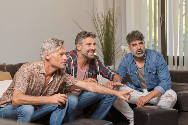 Männliche Freunde mittleren Alters zeigen weg und sitzen auf dem Sofa, während sie sich Streichhölzer ansehen und gemeinsam im Wohnzimmer Bier trinken - Foto, Bild