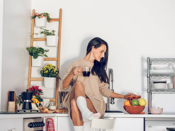 Graceful nuori etninen nainen pitkät tummat hiukset rento vaatteet juominen kuppi kahvia ja syö tuoreita hedelmiä istuessaan pöydällä keittiössä aamulla - Valokuva, kuva