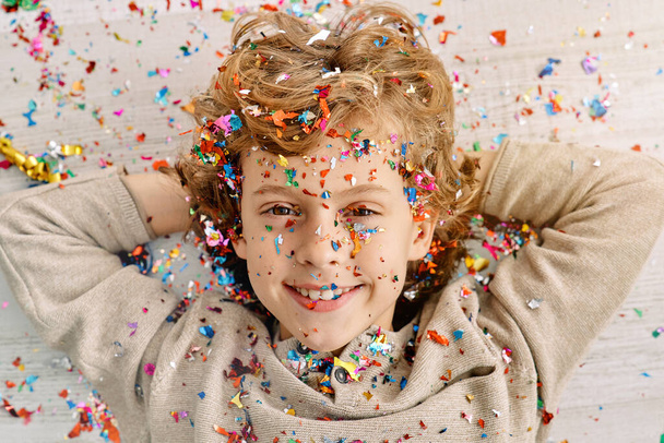 Little delightful child with curly fair hair lying with colorful confetti and looking at camera - Photo, Image