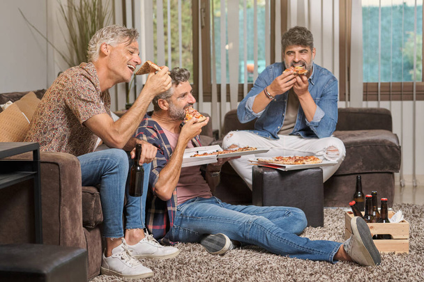 Grupo de amigos maduros do sexo masculino comer pizza saborosa e beber cerveja enquanto sentado na sala de estar em casa - Foto, Imagem