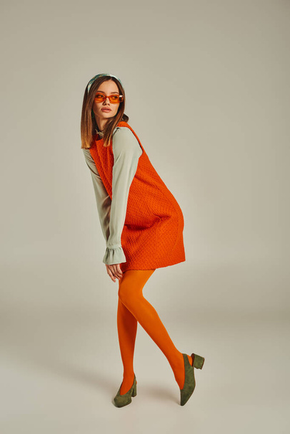 full length of vintage style woman in orange dress, tights and sunglasses looking away on grey - Photo, Image