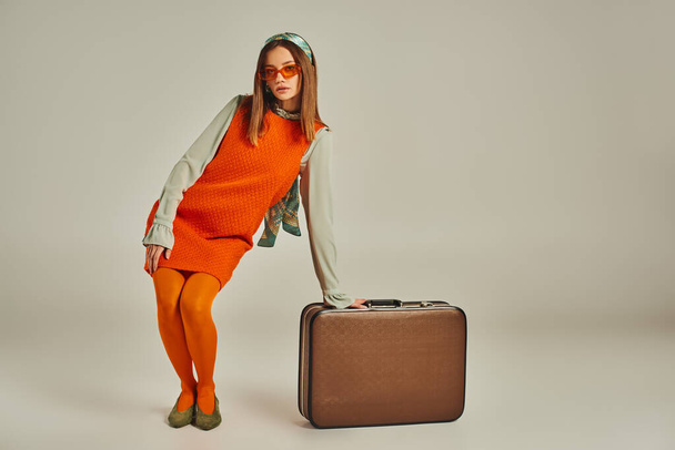 full length of glamour woman in orange dress posing near vintage suitcase on grey, fashion from past - Zdjęcie, obraz