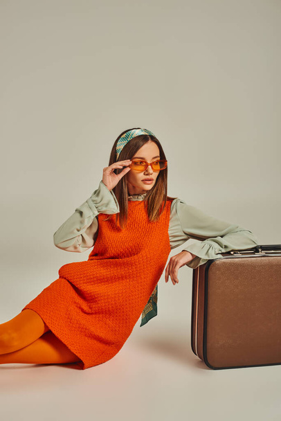 stylish woman in orange dress and sunglasses sitting near vintage suitcase and looking away on grey - Photo, Image