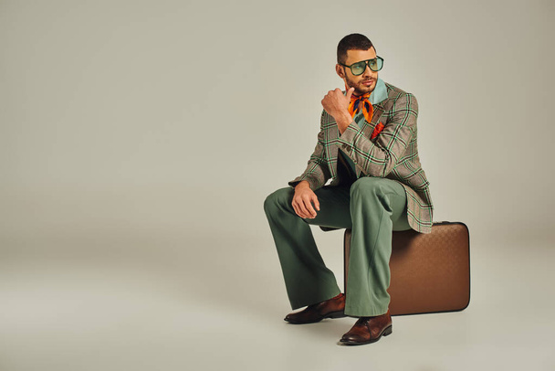 вдумчивый винтажный человек в солнцезащитных очках сидит на ретро чемодане и смотрит на серый - Фото, изображение