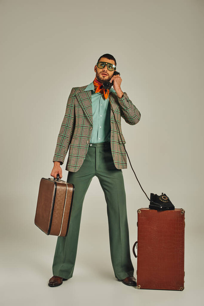 Stilvoller Mann in altmodischer Kleidung hält Vintage-Koffer in der Hand und telefoniert auf Smartphone in grau - Foto, Bild