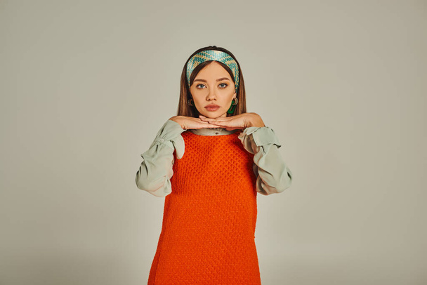young woman in colorful headband and orange dress posing with hands under chin on grey, retro style - Photo, Image