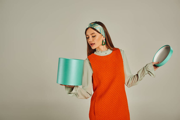 curious woman in orange dress and headband looking in gift box on grey, retro fashion photography - Foto, Bild