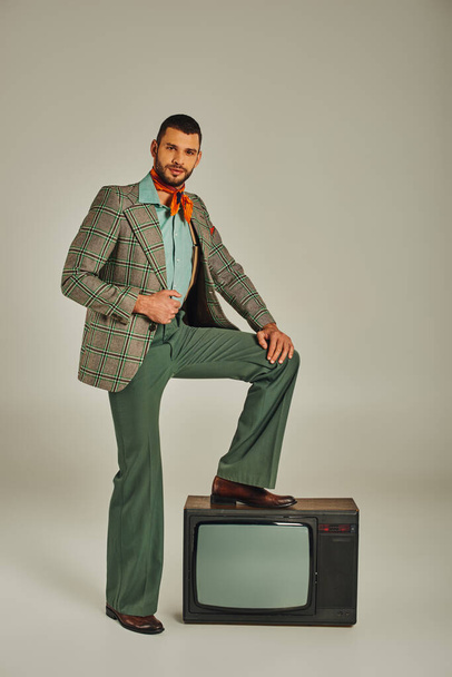 homem confiante em roupas retro elegantes pisando no aparelho de tv vintage e olhando para a câmera no cinza - Foto, Imagem