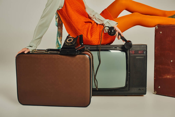 przycięty widok kobiety w pomarańczowej sukience siedzącej na zabytkowej walizce i TV na szarym, retro stylu - Zdjęcie, obraz
