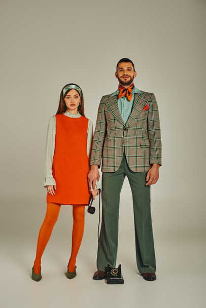 full length of young couple in trendy retro attire standing with handset f vintage phone on grey - Foto, Imagem
