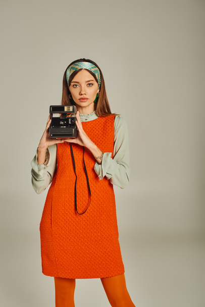 trendy woman in orange and colorful headband holding vintage camera on grey, retro-inspired style - Photo, Image