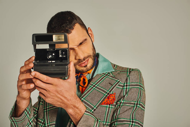 мужчина в клетчатом пиджаке и с красочным платком, фотографирующим на винтажную камеру в серой, ретро-моде - Фото, изображение