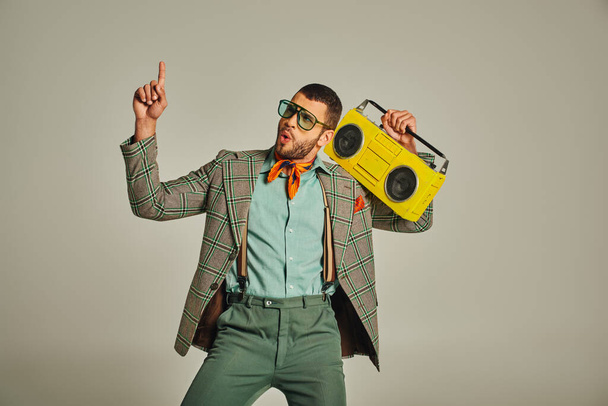 uomo eccitato in occhiali da sole in possesso di boombox giallo, indicando e ballando in grigio, stile vintage - Foto, immagini