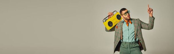 excited stylish man in sunglasses holding vintage boombox, pointing up and dancing on grey, banner - Photo, Image