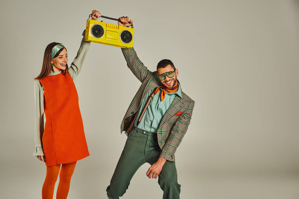 vzrušený pár ve stylovém vintage oblečení drží žlutý boombox a tanec na šedém pozadí - Fotografie, Obrázek