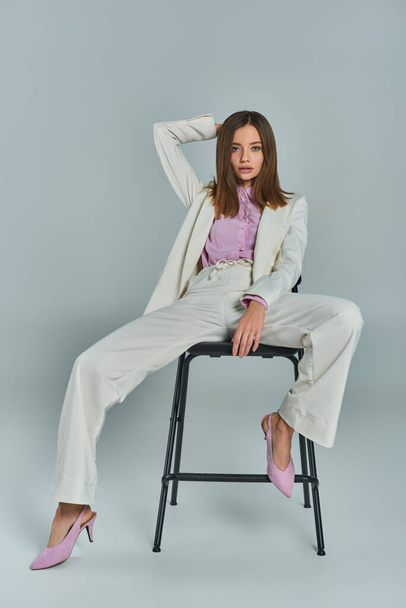 stijlvolle vrouw in wit elegant pak poseren op stoel en kijken naar de camera op grijs, volledige lengte - Foto, afbeelding