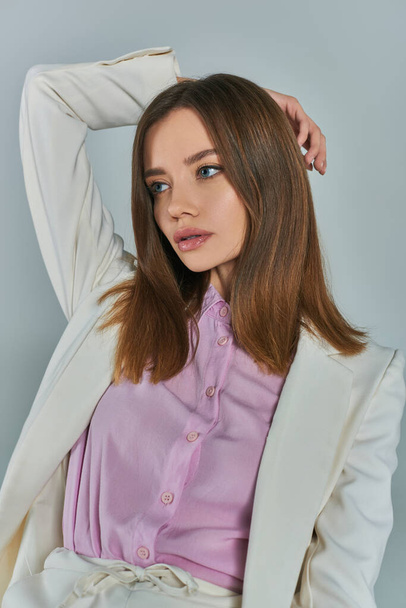 young charming woman in white blazer looking away with hand behind head on grey, fashion and style - Fotografie, Obrázek