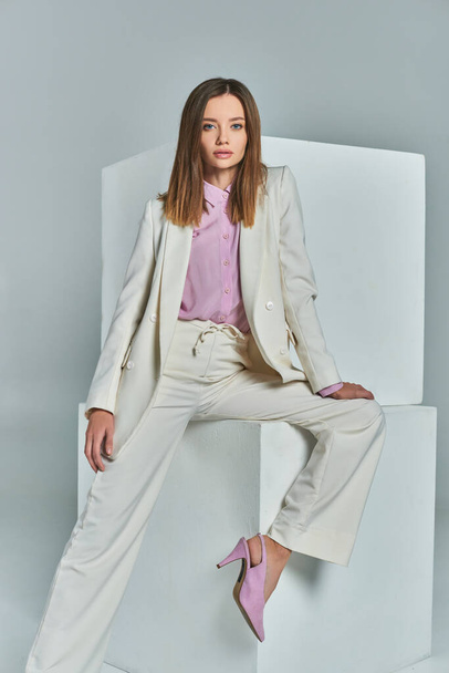 aantrekkelijke vrouw in elegante pak poseren in de buurt van witte kubussen op grijze, minimalistische zakelijke mode - Foto, afbeelding