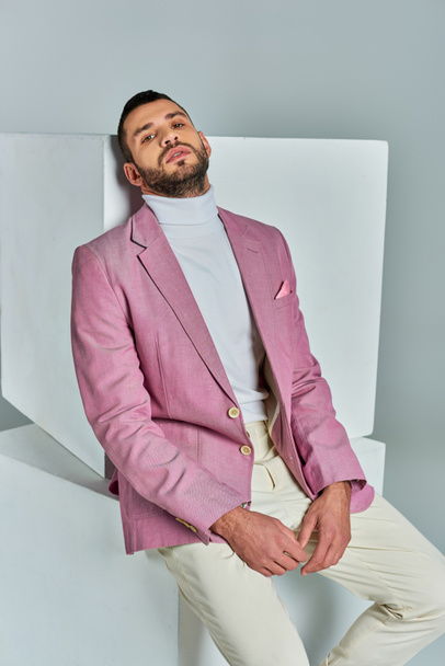 hombre carismático en blazer lila de moda sentado en cubo blanco y mirando a la cámara en gris - Foto, imagen