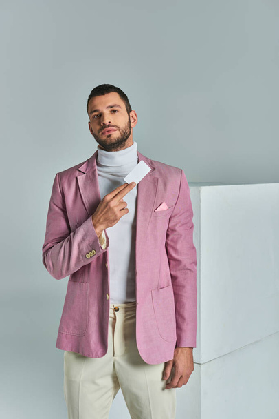 confiado hombre de negocios elegante en blazer lila celebración de la tarjeta de visita en blanco cerca de cubos blancos en gris - Foto, Imagen