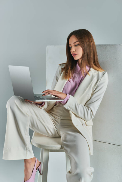young woman in elegant suit networking on laptop while sitting on stool near white cubes on grey - Photo, Image