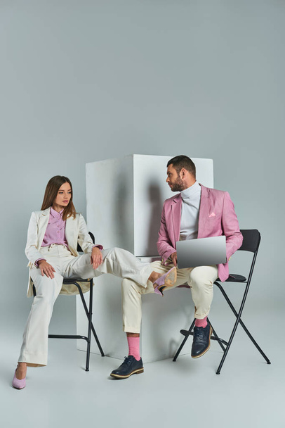 stylish man with laptop and young woman in white suit sitting on chairs near cubes on grey backdrop - Foto, Bild