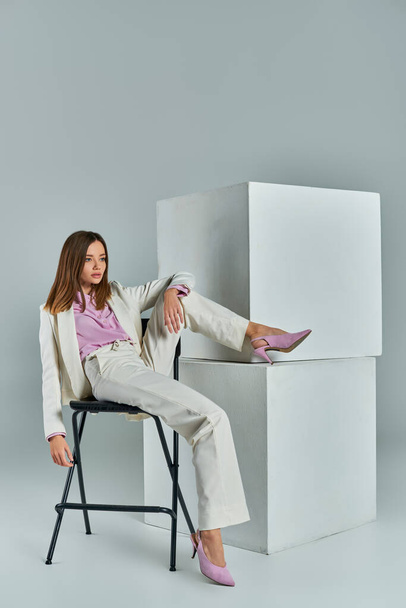 volledige lengte van charmante zakenvrouw in wit pak zitten op stoel in de buurt van witte kubussen op grijs - Foto, afbeelding