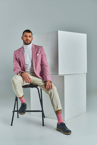 elegante man in zakelijke kleding zittend op stoel en kijkend naar camera in de buurt van witte kubussen op grijs - Foto, afbeelding