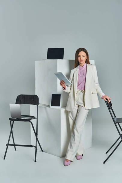 junge Frau im weißen Anzug mit Laptop in der Nähe von Stühlen und digitalen Geräten auf weißen Würfeln auf grau - Foto, Bild