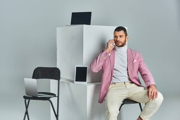 впевнений стильний чоловік говорить на смартфоні, сидячи біля пристроїв на білих кубиках на сірому - Фото, зображення