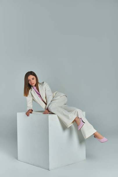 longitud completa de la mujer joven en traje elegante posando en cubo blanco sobre fondo gris en el estudio - Foto, imagen