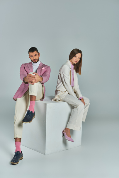 módní obchodní pár v elegantním formální oblečení sedí na bílé kostce a dívá se na kameru na šedé - Fotografie, Obrázek