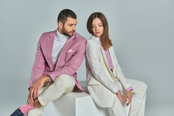 young and fashionable couple in elegant suits sitting on white cube on grey backdrop in studio - Photo, Image
