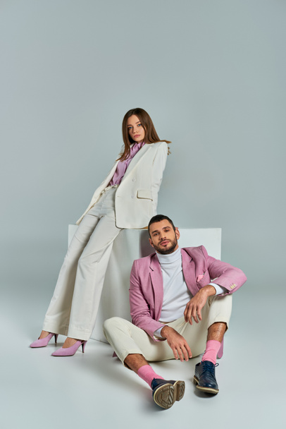 man in lila blazer zitten op de vloer in de buurt van vrouw in stijlvolle pak poseren in de buurt van witte kubus op grijs - Foto, afbeelding