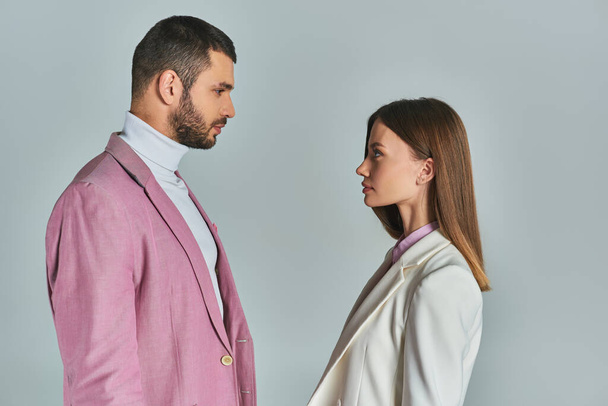 side view of young couple in stylish blazers looking at each other on grey, minimalistic fashion - Photo, Image