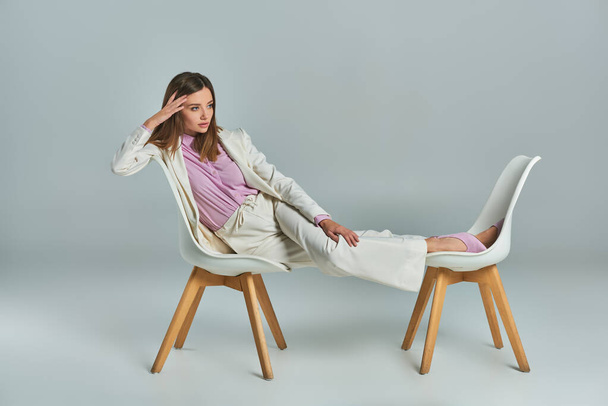 молодая и задумчивая деловая женщина в белом элегантном костюме позирует на креслах и смотрит в сторону серого - Фото, изображение